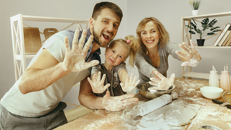 Cute Little Girl And Her Beautiful Parents Preparing The Dough For The Cake In Kitchen At Home