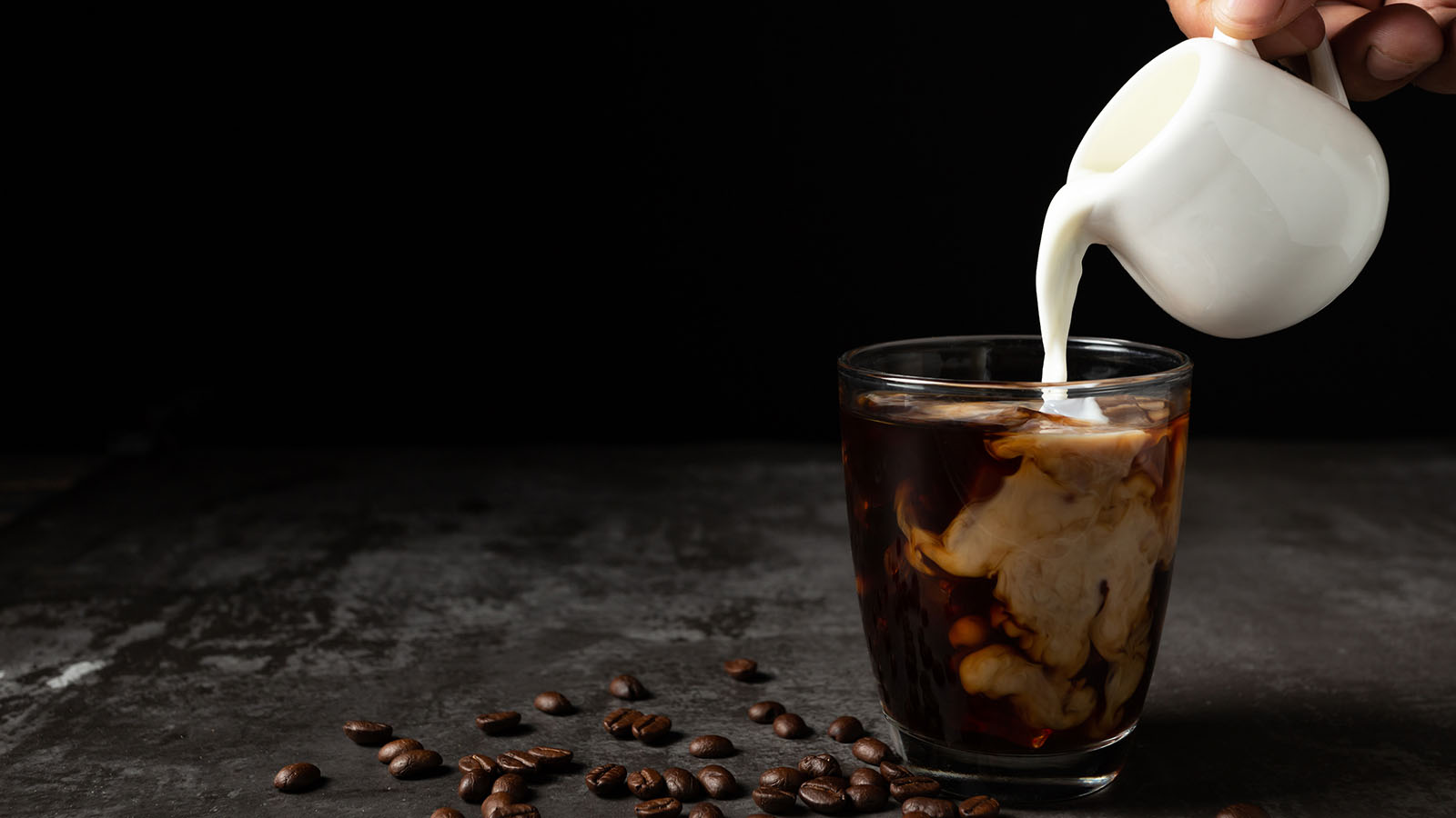 Milk Pouring Into Iced Black Coffee On Table And Black Backgroun